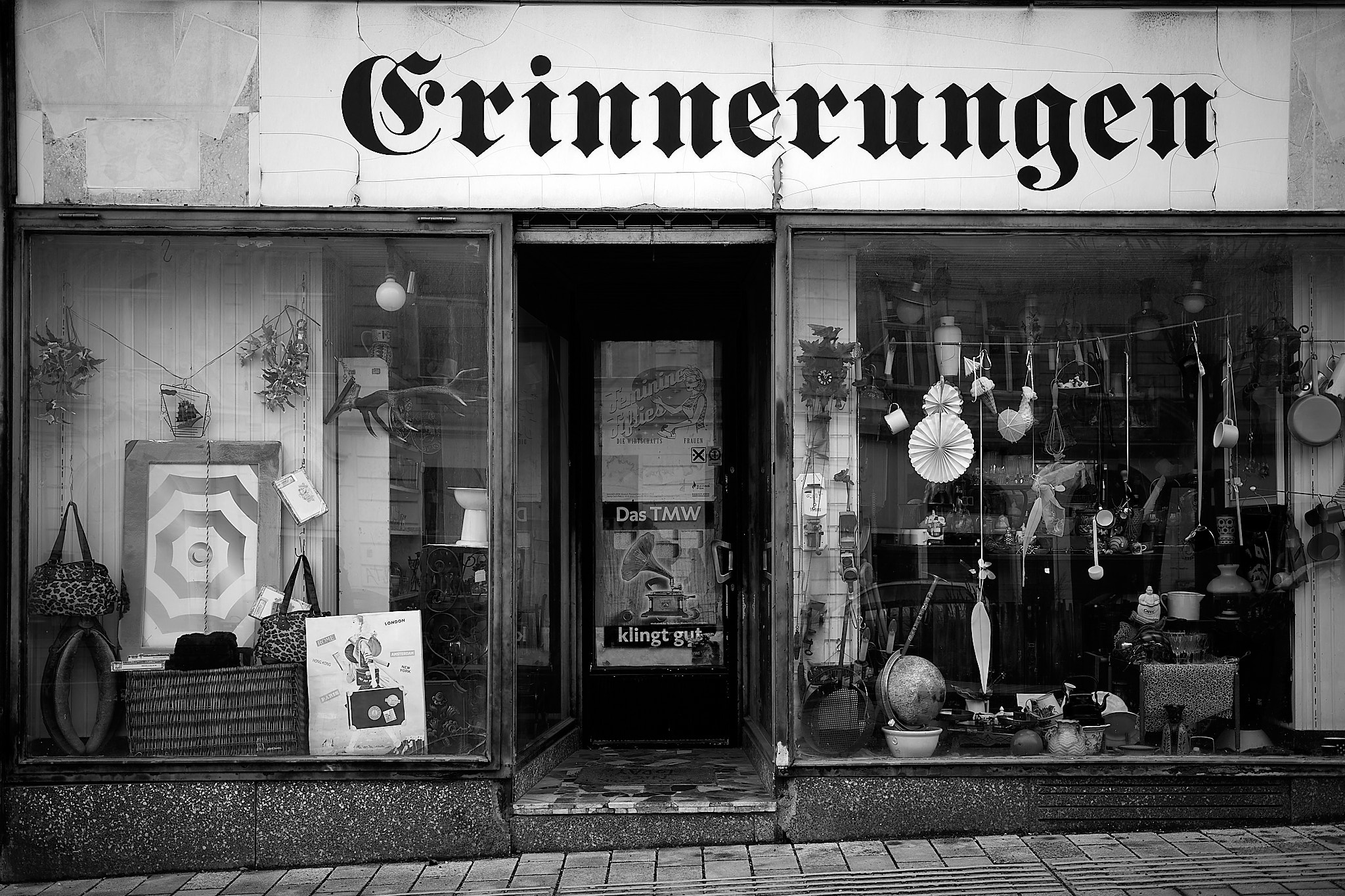 Black and white photo of a quaint storefront with the word "Erinnerungen" above the window, displaying various items including bags, lamps, ornaments, and a poster with text "Das TMW klingt gut" visible through the glass door.